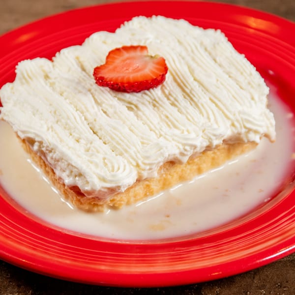 A slice of tres leches cake with whipped cream and a strawberry on a red plate.